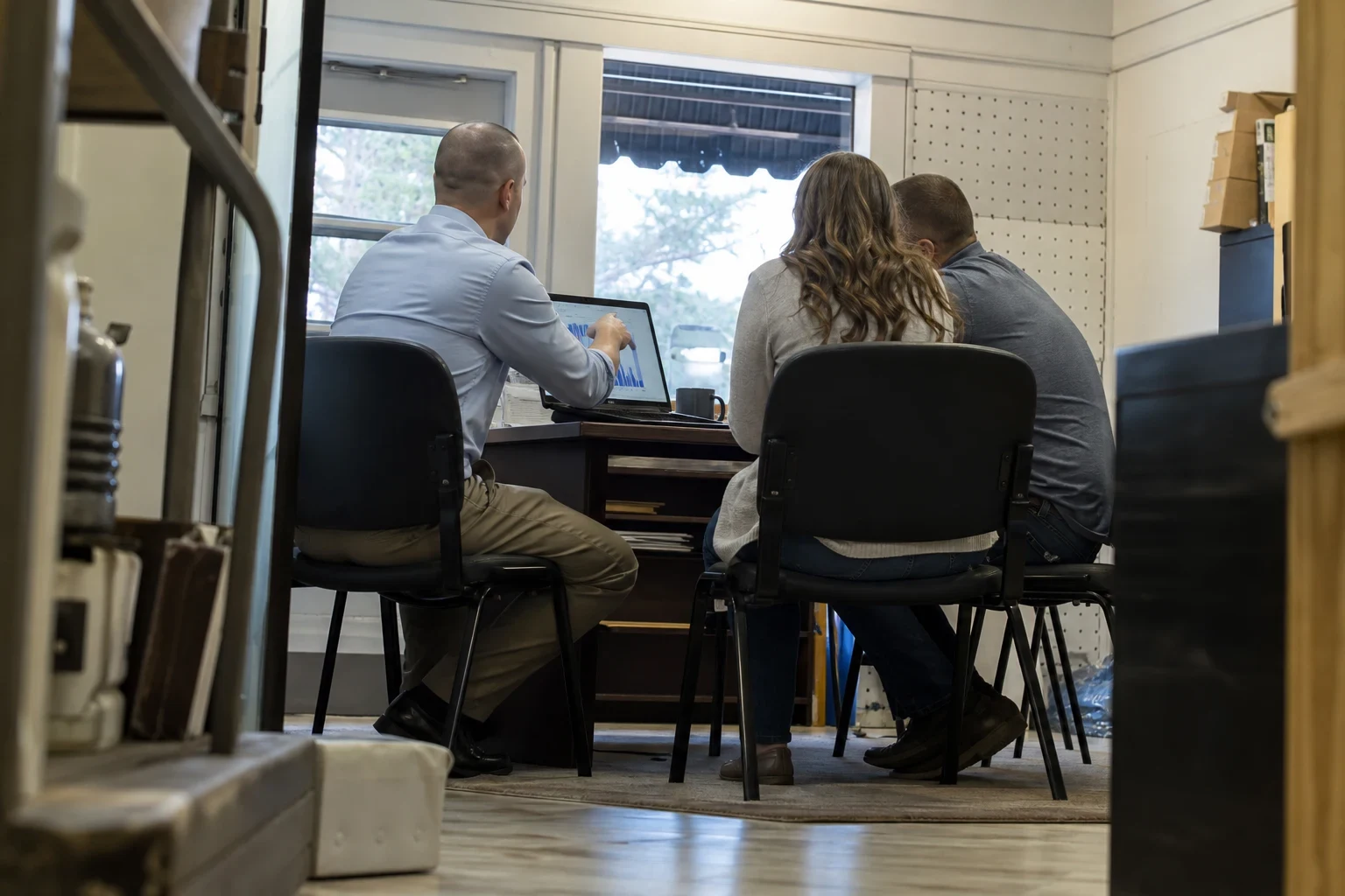 Man in a light blue button-down shows a chart on a laptop to a couple seated beside him at a desk in a small storefront office with pegboard walls and a window.